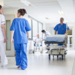 biały korytarz szpitalny, w tle personel medyczny, dwie kobiety tyłem w niebieskim i białym fartuchu, z naprzeciwka łózko szpitalne pchane przez mężczyzne w niebieskim fartuchu